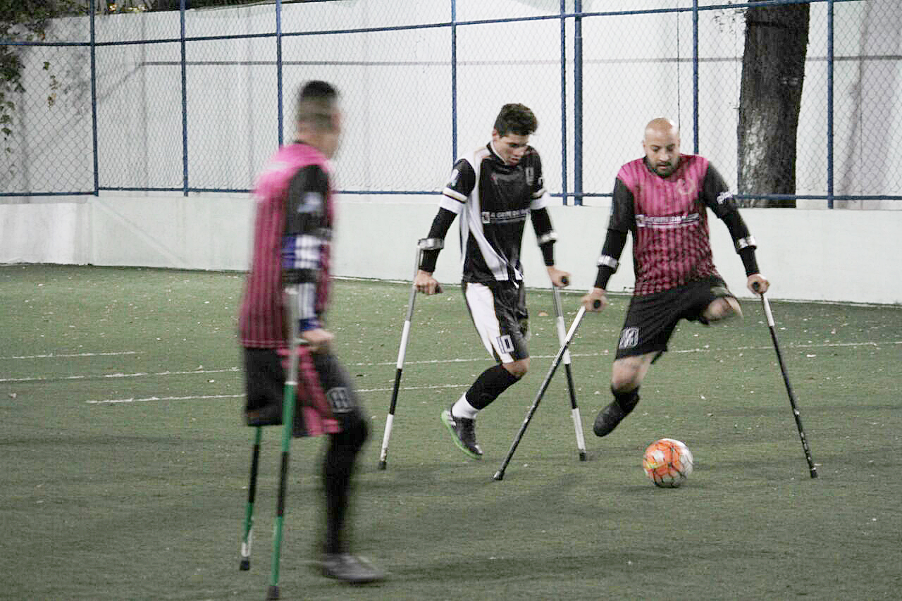 Jogadores amputados jogam bola durante competição. Eles estão no campo de futebol e disputam a bola.